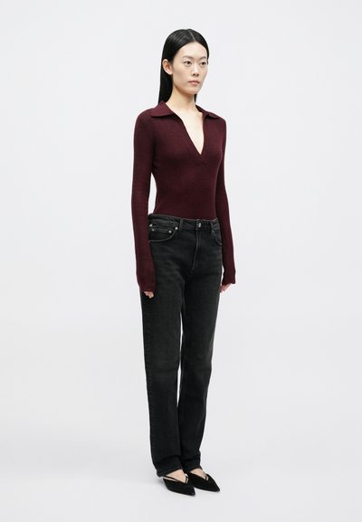 Tmavohnedý pletený top s dlhým rukávom a hlbokým výstrihom do V, skombinovaný s čiernymi džínsami s rovnými nohavicami. Modelka má na nohách čierne baleríny s uhlovým dizajnom.