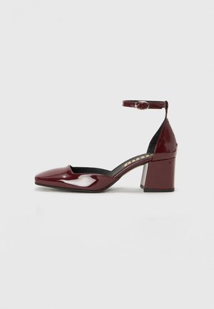 Burgundy patent leather high heel shoe with a pointed toe, ankle strap, and chunky heel. Smooth texture and black interior lining.