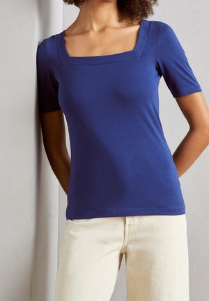 Femme portant un haut bleu ajusté à manches courtes avec un décolleté carré et un pantalon beige clair, debout les mains derrière le dos.