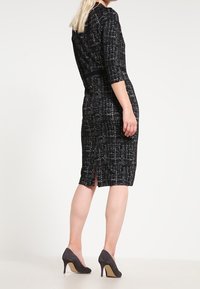 Svart och vit texturerad klänning med trekvartslånga ärmar, knälång design och en sprund i ryggen, kombinerad med grå högklackade skor.