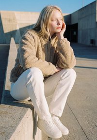 Młoda kobieta w beżowej polarowej kurtce i białych spodniach siedzi na betonowej krawędzi, opierając głowę na dłoni, zapatrzona zamyślona w światło słoneczne.