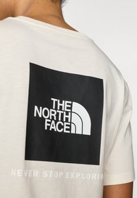 Βαμβακερό t-shirt σε εκρού χρώμα με μεγάλο μαύρο τετράγωνο που φιλοξενεί λευκό λογότυπο στο πίσω μέρο ς. Το κείμενο από κάτω γράφει "ΠΟΤΕ ΜΗΝ ΣΤΑΜΑΤΑΣ ΝΑ ΕΞΕΡΕΥΝΑΣ" λευκά.