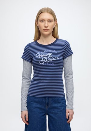 Jeune femme aux cheveux blonds et raides porte un t-shirt à manches courtes bleu et marine rayé, superposé à un top gris à manches longues et un jean bleu.