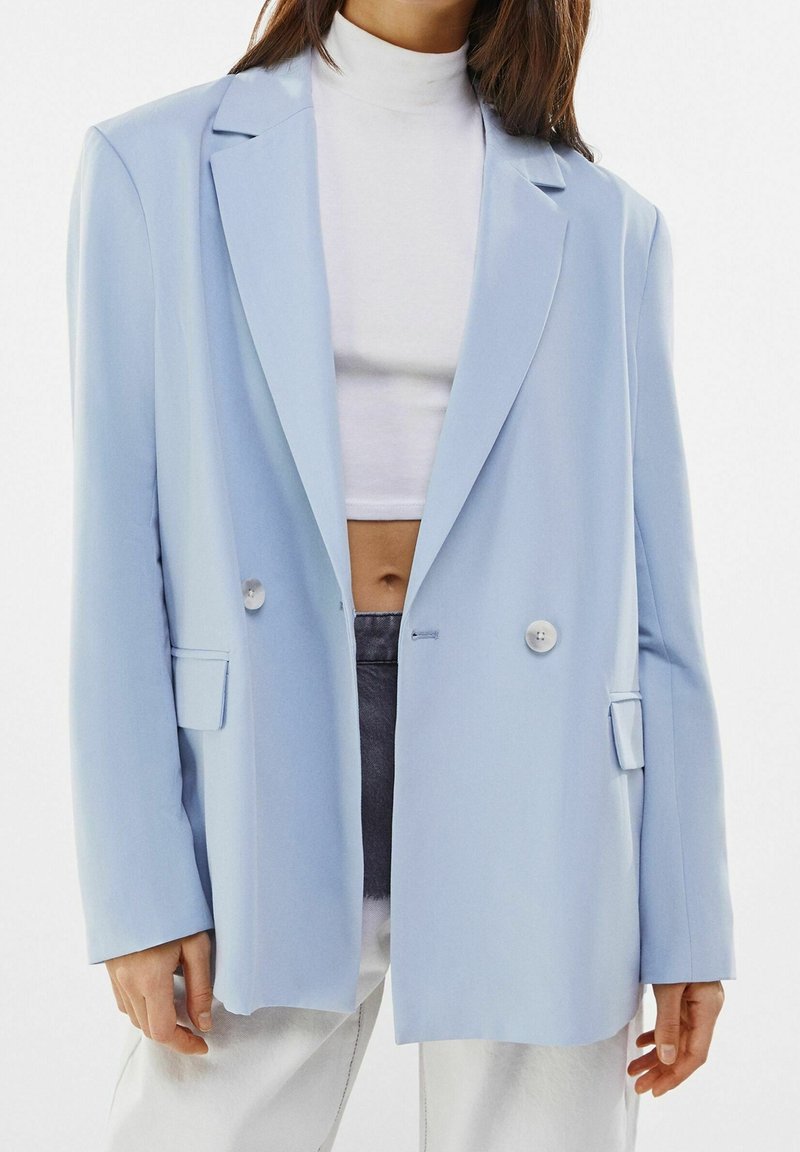 Light blue blazer with a loose fit, notch lapels, double buttons, and side pockets. Smooth fabric, paired with a white turtleneck underneath.