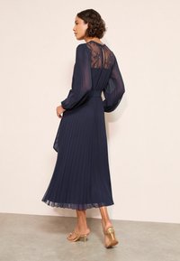 Granatowa sukienka plisowana z długimi przezroczystymi rękawami i koronkowym dekoltem. Projekt cechuje się paskiem w talii oraz zwiewną sylwetką w kształcie litery A.