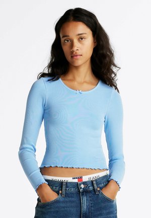 Helles blaues, geripptes Langarmoberteil mit rundem Halsausschnitt und gewelltem Saum. Trägt dazu hoch taillierte Jeans mit einem Logo-Bund.