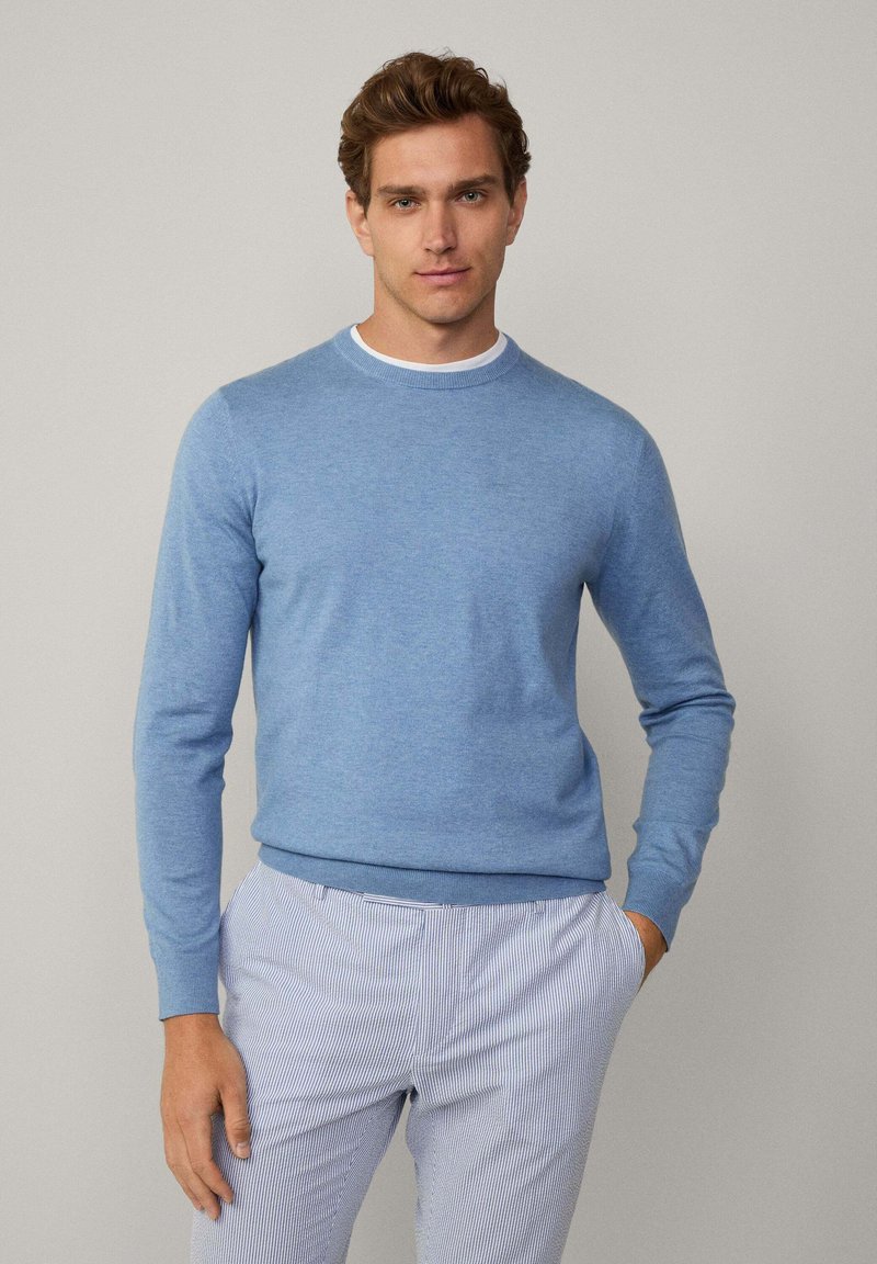 Jasnoniebieski sweter dziany z okrągłym dekoltem i długimi rękawami, zestawiony z jasnymi spodniami w paski. Gładka struktura, luźny krój.