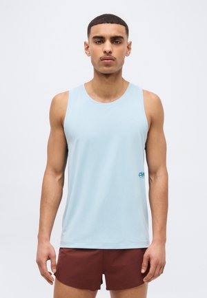 Νέος άντρας με κοντά μαλλιά που φοράει ανοιχτό μπλε αμάνικο αθλητικό τοπ και μπορντό σορτς, στέκεται μπροστά σε απλό φόντο.