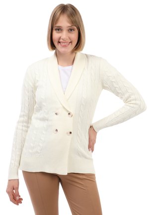 Krémový pletený cardigan s šálovým límcem, dvojitými knoflíky a žebrovanými manžetami, kombinovaný s úzkými béžovými kalhotami.