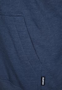 Mörkblå sweatshirt i mjukt tyg, med en framficka och en liten svart "Burton"-etikett på sidan. Slät textur och avslappnad design.