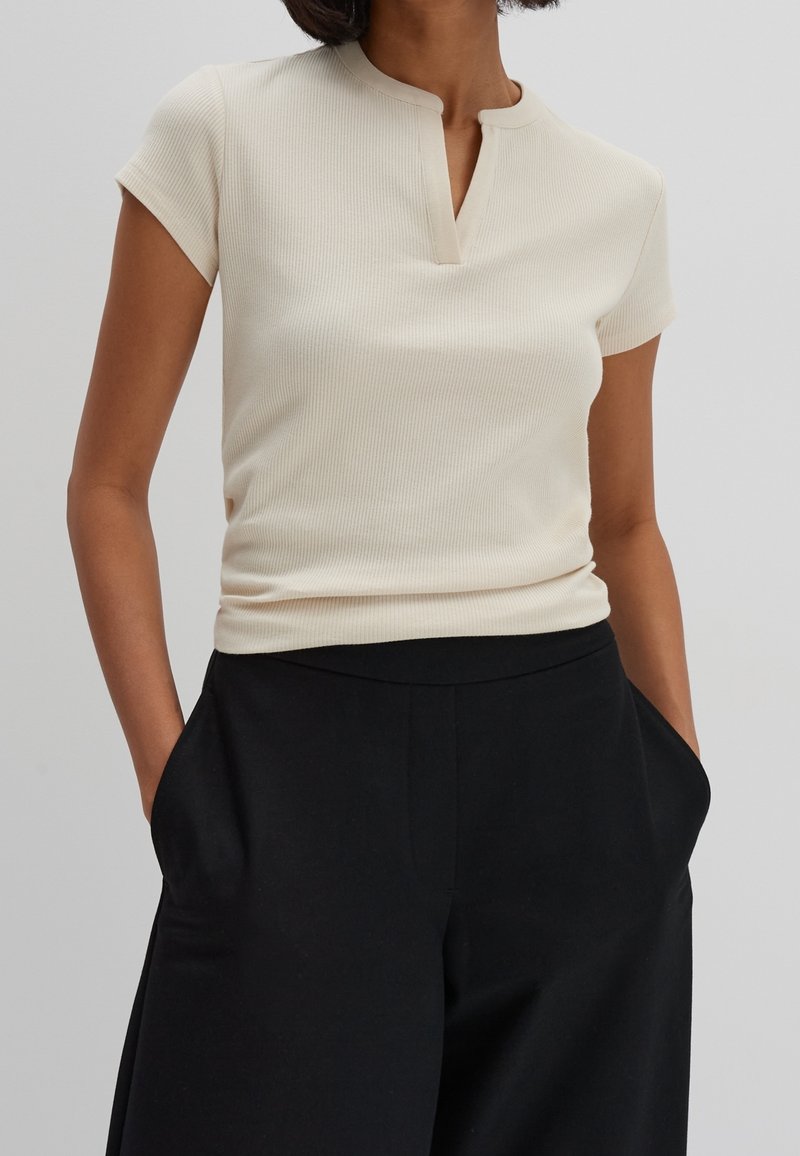 Kremowy, prążkowany top z krótkim rękawem i kołnierzykiem mandaryńskim, w połączeniu z czarnymi spodniami o wysokim stanie z bocznymi kieszeniami. Gładka faktura.