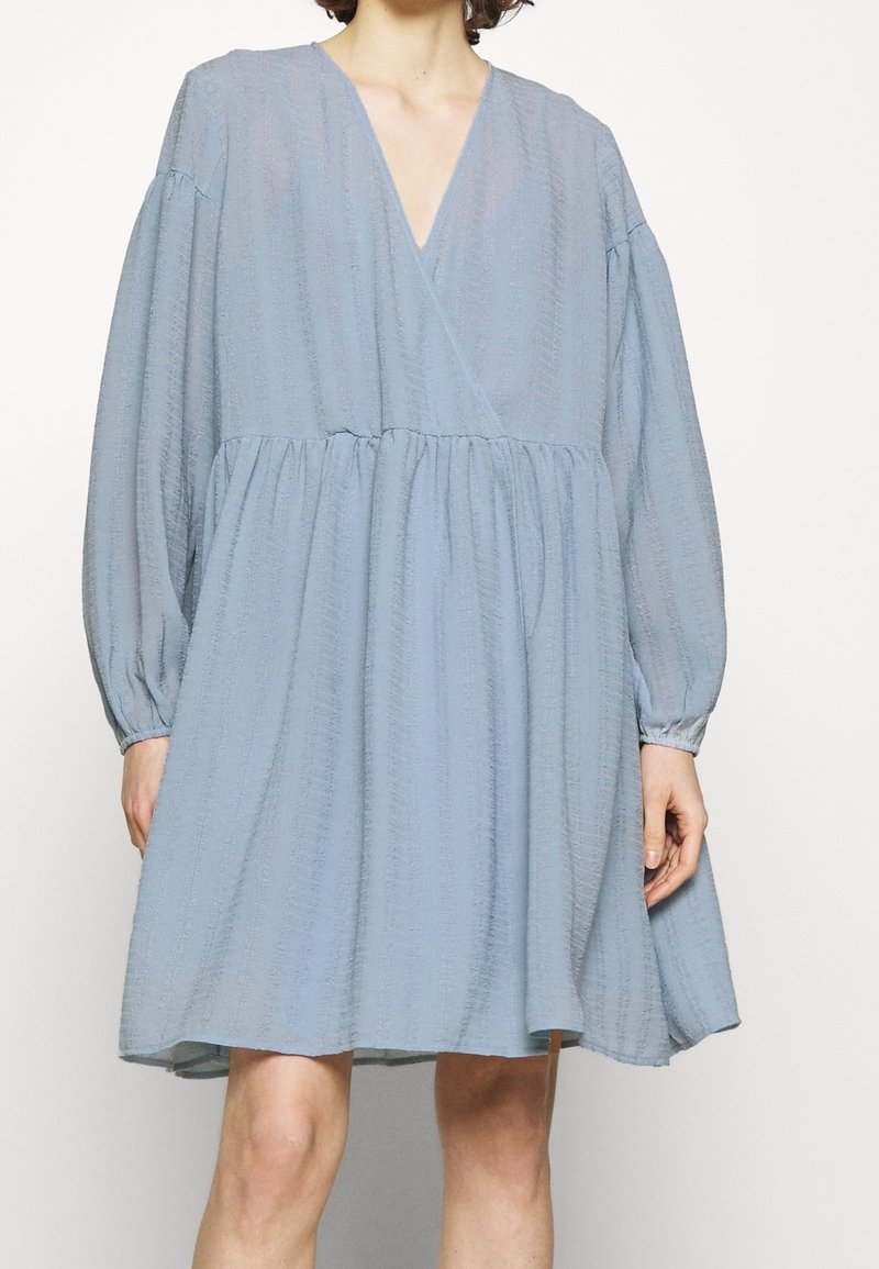 Femme portant une robe ample texturée bleu clair avec de longues manches bouffantes et un col en V, tombant au-dessus des genoux.