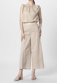Beige volná blůza s šňůrkovým výstřihem, kombinovaná s širokými béžovými kalhotami s švem. Model má na sobě odpovídající skluzavkové sandály.