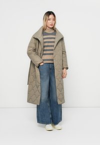 Lange, gewatteerde, lichtbeige pufferjas met een grote kraag, knoopsluiting, gecombineerd met een gebreide trui met horizontale strepen en een wijdvallende spijkerbroek.