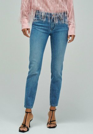 Mujer que lleva unos vaqueros ajustados de color azul claro, tacones altos negros con tiras y una blusa rosa de manga larga con detalles de flecos en las mangas y el dobladillo.