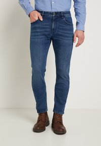 Mörkblå denimjeans med slim fit, med sydda fickor, subtila blekningar och slitna kanter vid fållarna, kombinerade med bruna stövlar.
