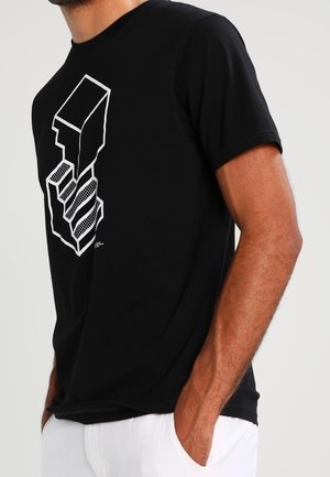 Mann trägt ein schwarzes T-Shirt mit weißem abstraktem geometrischem Design und weiße Hosen, Hände in den Taschen, vor neutralem Hintergrund.