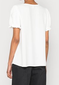 Biała bluzka z krótkimi bufiastymi rękawami, wykonana z miękkiego materiału, z okrągłym dekoltem i luźnym krojem. Posiada delikatne falbany na krawędziach rękawów.