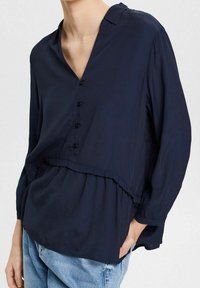 Blouse boutonnée bleu marine avec une coupe décontractée, un décolleté en V, des manches longues et un ourlet à volants. Confectionnée dans un tissu léger et lisse.