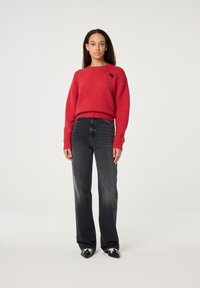 Roter Wollpullover mit Rundhalsausschnitt und Logodetail, kombiniert mit hochgeschnittenen schwarzen Jeans. Das Model steht vor einem neutralen Hintergrund.