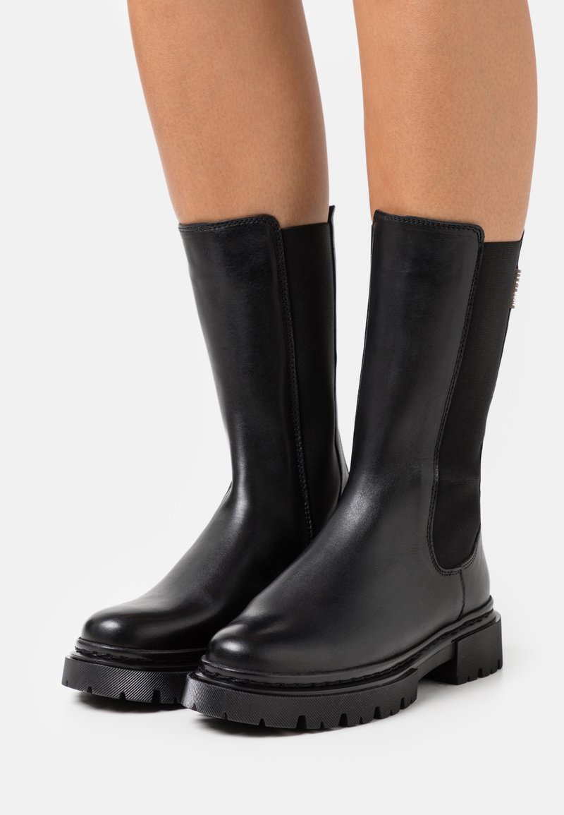 Bottes en cuir noires mi-mollet avec semelle épaisse et panneaux latéraux élastiques portées sur jambes nues sur fond blanc.
