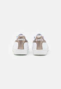 Baskets blanches avec un dessus lisse, un accent de talon doré métallique et une semelle intermédiaire texturée blanche. La marque sur le talon présente un logo.