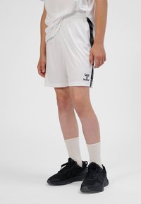 Weiße Sportshorts mit schwarzen Fischgrät-Seitenstreifen und Logo. Kombiniert mit schwarzen Turnschuhen und cremefarbenen Knöchelsocken.