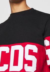 Felpa nera e rossa con grandi lettere bianche "COS" sulla parte del petto. Presenta un collo rotondo e un tessuto di cotone morbido.