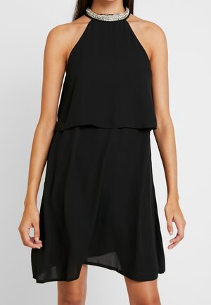 Femme portant une robe noire sans manches à col halter avec un col orné de perles argentées, un corsage superposé et une jupe fluide longueur genou.