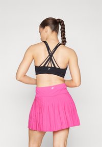 Sujetador deportivo negro con tirantes entrecruzados; combinado con una falda corta rosa con plisado, que presenta una cinturilla ancha y un detalle de logo en la parte frontal.