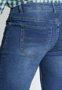 Jeans de mezclilla azul con un ajuste ceñido, que cuentan con un diseño clásico de cinco bolsillos y costuras en contraste. Textura suave y acabado ligeramente desgastado.