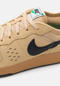 Baskets en suede et mesh beige avec un swoosh noir de Nike, des lacets beiges et une semelle en caoutchouc. Elles présentent des perforations pour la ventilation et un intérieur vert.