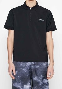 Černé polo tričko s krátkým rukávem, se zipovým límcem a kapsou na hrudi, vyrobené z hladké tkaniny s jemným leskem, s malým logem.