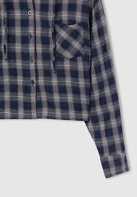 Camicia a scacchi blu navy e grigia con maniche lunghe e taschino sul petto, su uno sfondo chiaro.