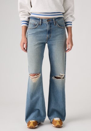 Femme portant un jean flare déchiré bleu clair, un pull blanc à rayures bleues aux poignets et à l'ourlet, et des baskets marron avec des lacets blancs.
