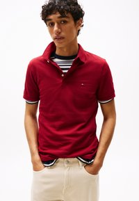 Rdeča polo majica s kratkimi rokavi in ovratnikom, z majhnim logotipom na prsih. Nošena čez črtasto majico s kratkimi rokavi. Hlače nevtralne barve.