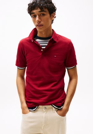 Polo rossa a maniche corte con colletto, con un piccolo logo sul petto. Indossata sopra una maglietta a maniche corte a righe. Pantaloni di colore neutro.