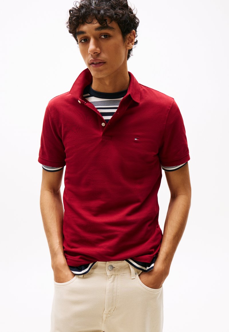 Rdeča polo majica s kratkimi rokavi in ovratnikom, z majhnim logotipom na prsih. Nošena čez črtasto majico s kratkimi rokavi. Hlače nevtralne barve.