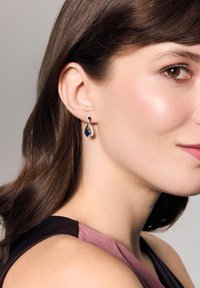 Srebrne kolczyki w kształcie łzy z niebieskim kamieniem szlachetnym i przejrzystymi akcentami, zawieszone na zakrzywionym wzorze.