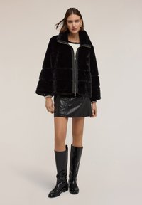Kobieta w czarnej kurtce z sztucznego futra na zamek, czarnej skórzanej mini spódnicy i błyszczących czarnych długich butach, stojąca na jasnym tle.