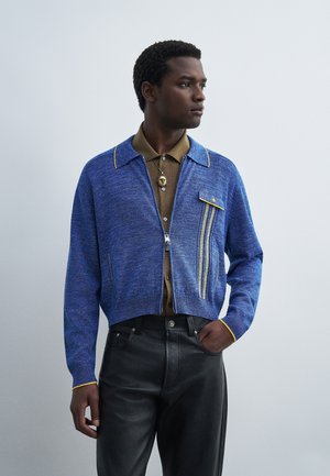 Homme portant un cardigan bleu zippé avec une bordure jaune sur un polo marron et un pantalon en cuir noir, regardant vers la droite avec une main dans la poche.
