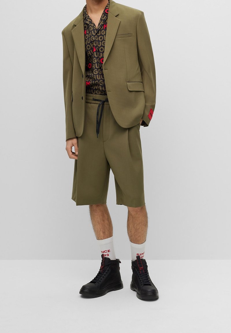 Homme portant un blazer vert olive et un short assorti jusqu'aux genoux, une chemise noire à motifs, des chaussettes blanches avec un texte rouge, et des chaussures montantes noires.