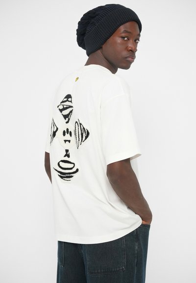 Λευκό βαμβακερό t-shirt με μαύρο αφηρημένο σχέδιο στην πλάτη. Το μοντέλο φοράει ναυτικό πλεκτό σκουφάκι και σκούρα παντελόνια. Απλή και μοντέρνα γραμμή.