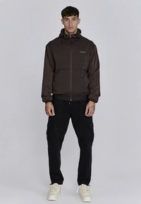 Brun jacka med dragkedja och mönstrade ärmar, bärs med svarta cargobyxor och krämfärgade skor. Har huva och logotyp på bröstet.