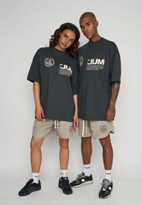 Dos individuos vestidos con camisetas de gran tamaño en gris oscuro con logotipos impresos, combinadas con pantalones cortos beige. Zapatillas en tonos negros y grises.