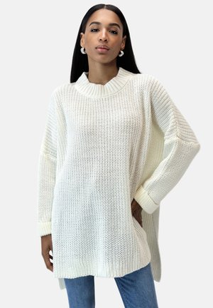 Maglione over-size color crema con una texture a coste e un colletto alto ripiegato. Presenta spacchi laterali e maniche ampie, abbinato a jeans di denim.