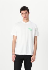 Valkoinen puuvillapaitajoukkue, jossa on lyhyet hihat ja pyöreä pääntie, vasemmassa rintataskussa vihreä logo. Yksinkertainen, koristamaton muotoilu.