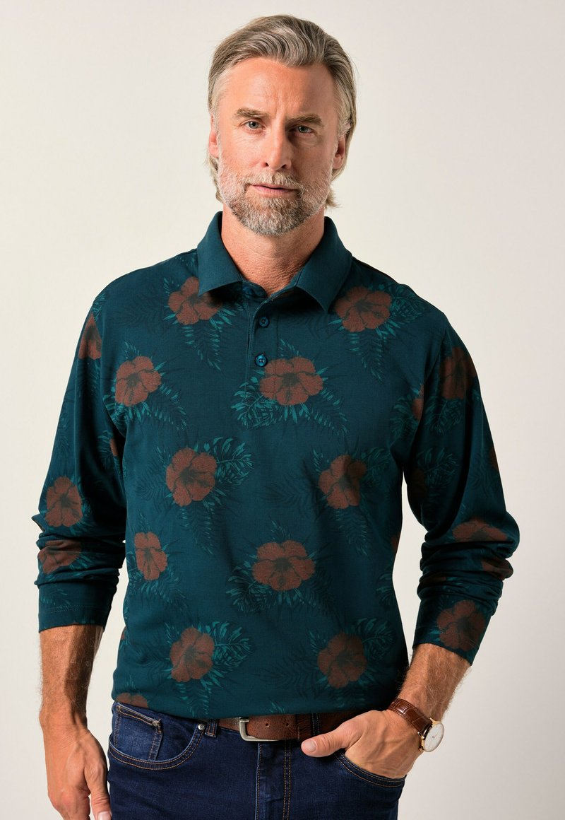 En medelålders man med grått hår och skägg bär en mörk teal långärmad polo med bruna blommönster och blå jeans, med handen i fickan.