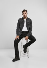 Sivi traper jakna s džepovima na prsima, bijela majica, crne hlače i crne čizme, sjedi na bijelom stolcu uz svijetlu pozadinu.