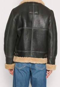 Achteraanzicht van een persoon in een zwarte leren jas met beige schapenwollen trim op de manchet en zoom, gecombineerd met een blauwe spijkerbroek.